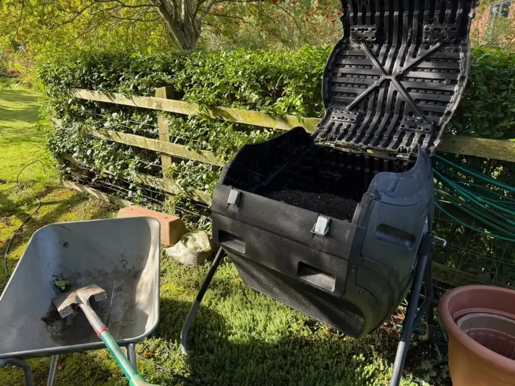 A compost tumbler standing open beside a wheelbarrow of finished compost