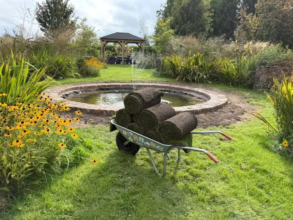 A wheelbarrow of turf rolls beside a formal pond, ready for laying a lawn around its edge