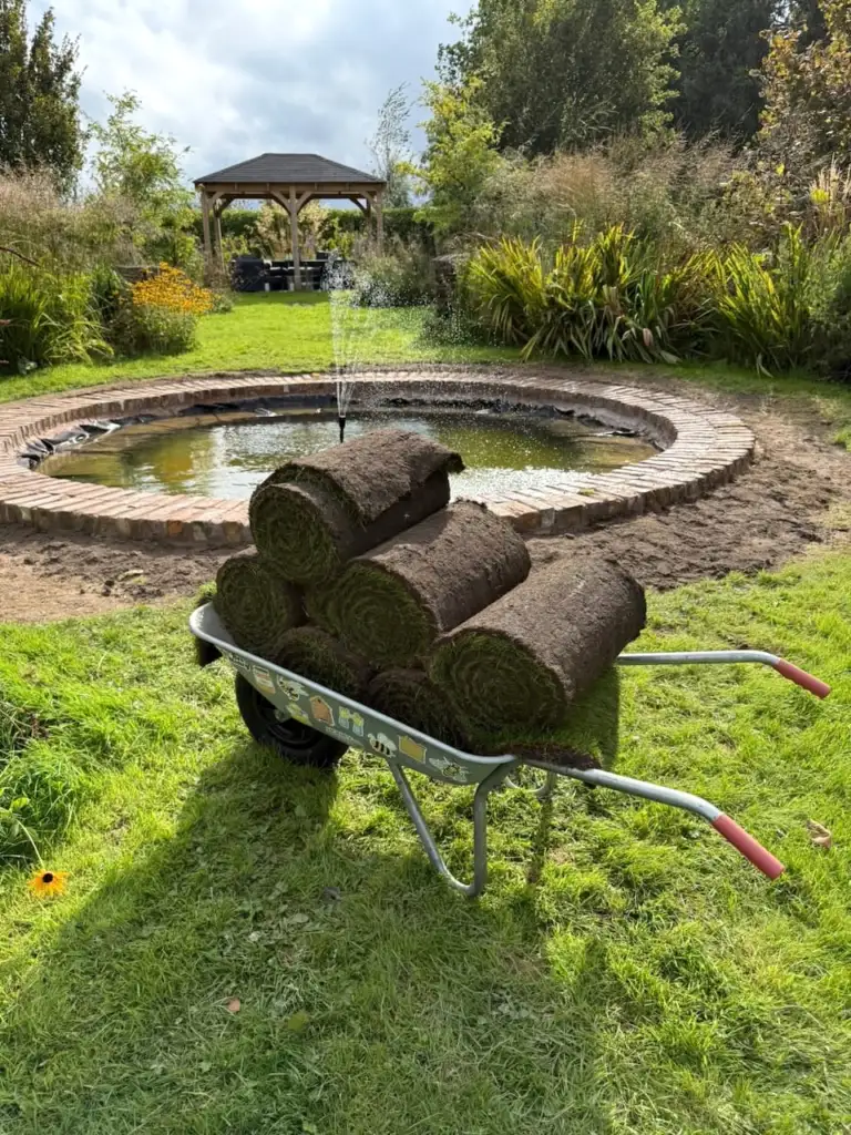 Rolls of freshly cut turf stacked in a wheelbarrow beside the stone edge of a newly fitted pond