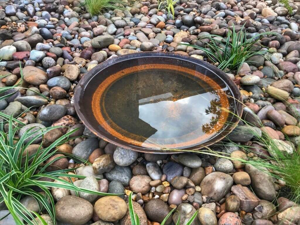 A corten steel water bowl on cobbles