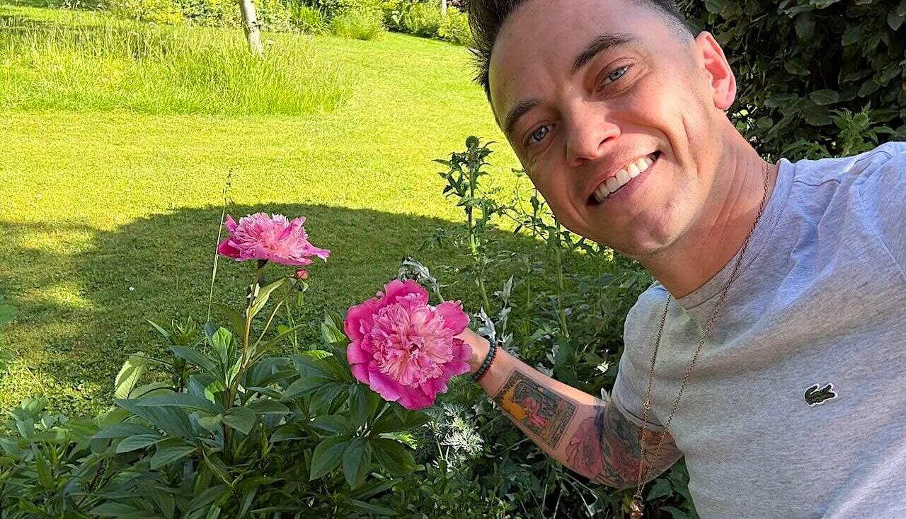 Lee Burkhill holding a bunch of pink Peonies beside a lawn and flower bed