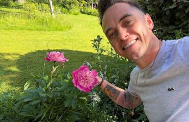 Lee Burkhill holding a bunch of pink Peonies beside a lawn and flower bed